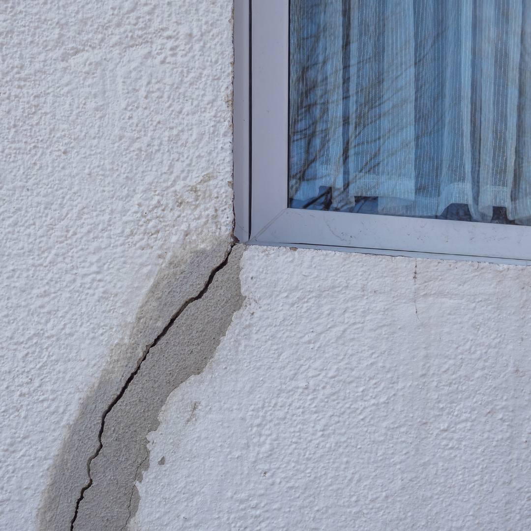 A close-up of a wall with a visible crack near a window, showing textured white paint and a gray window frame.