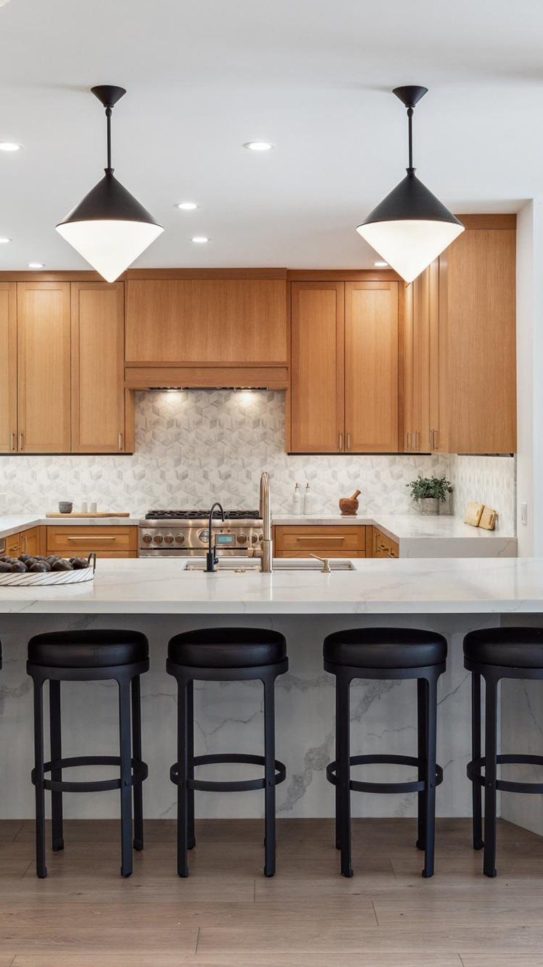A modern kitchen featuring wooden cabinets, marble countertops, black bar stools, and stylish pendant lighting.