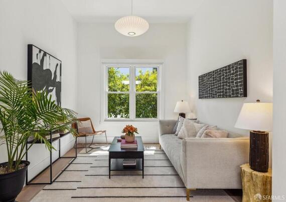 A bright living room featuring a light sofa, coffee table, and plants, with large windows and modern decor.