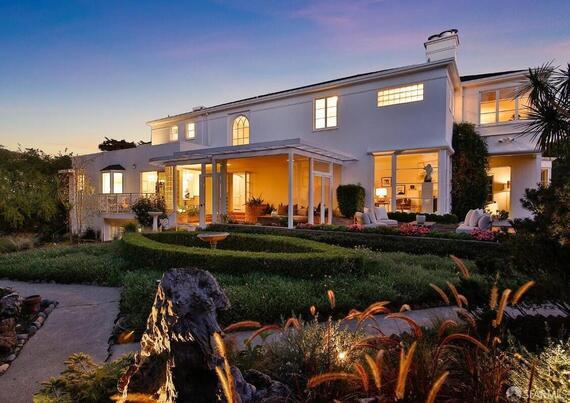 A spacious white house with a landscaped garden, illuminated at dusk, featuring a porch and lush greenery.